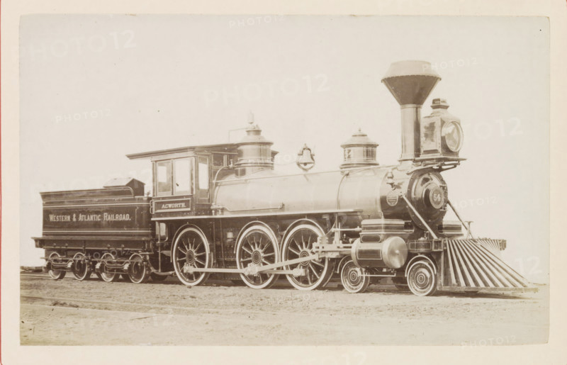 American freight steam locomotive, photograph, page, United States of America, c. 1871 - in or before 1881, photographer, North America, paper, albumen print, height 125 mm x width 198 mm, Photo on page 84., steam locomotive, A sepia-toned photograph of a vintage steam locomotive positioned on tracks, showing the engine and its attached coal car. The locomotive has a tall, flared smokestack and a large round headlamp mounted at the front above a slatted pilot. Two cylindrical domes sit along the top of the boiler, and a bell hangs between them. The cab has windows and a roof overhang, and the coal car behind it has a raised rear section. Large spoked driving wheels and smaller leading wheels are visible, with connecting rods and mechanical linkages along the side. The surface beneath the train appears to be dirt, and the background is a pale, empty sky. The image border shows a thin margin around the photograph.