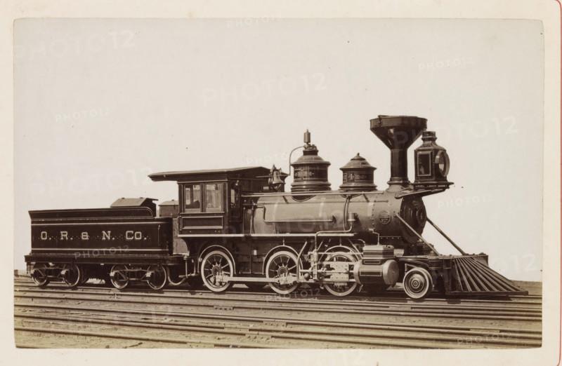 American freight steam locomotive, photograph, page, United States of America, c. 1871 - in or before 1881, photographer, North America, paper, albumen print, height 124 mm x width 198 mm, Photo on page 90., steam locomotive, A sepia-toned photograph of a vintage steam locomotive and its tender on parallel tracks, showing the side profile of the engine with a rounded smokebox, a tall flared smokestack, and two domed fittings on the boiler; the cab has rectangular windows and a roof overhang, and the tender sits behind with a sloped top. The visible running gear includes large spoked driving wheels, connecting rods, and various pipes and mechanical linkages along the side. At the front there is a metal pilot with slatted bars angling downward and a headlight housing mounted above the smokebox. The image shows the train centered horizontally against a pale background with the rails and sleepers beneath.