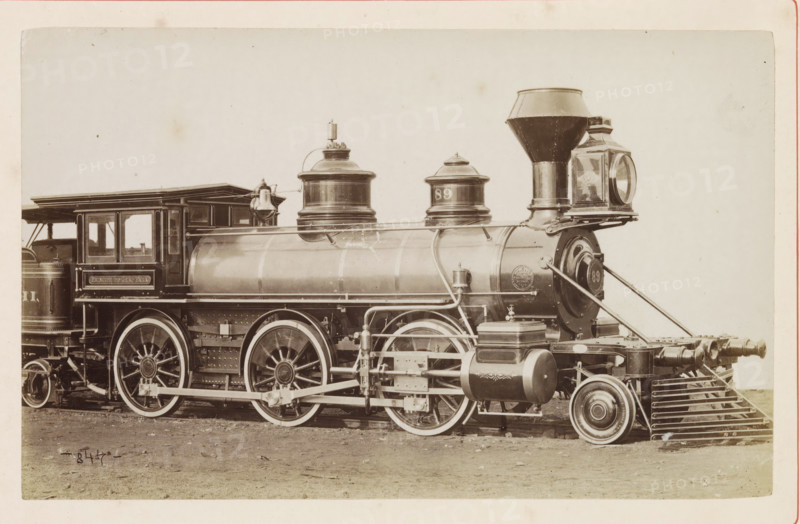American freight steam locomotive, photograph, page, number: '844', United States of America, c. 1871 - in or before 1881, photographer, North America, paper, albumen print, height 124 mm x width 198 mm, Photo on page 96., steam locomotive, A sepia-toned photograph of a vintage steam locomotive showing the side of the engine and part of the tender, with large spoked driving wheels and a visible cowcatcher at the front. The cylindrical boiler runs along the center with domed steam domes on top and a tall conical smokestack near the front. The cab has open windowed panels and a roof extending over the rear of the boiler. Brass fittings, pipes, and valves run along the boiler and frame, and a rectangular lantern with a round glass face is mounted at the front. Various mechanical linkages and rods connect the wheels, and there is a cylindrical tank and other rounded components attached near the pilot. The ground beneath the locomotive is bare and slightly uneven.