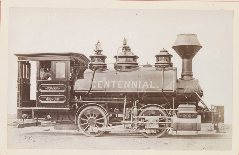 American steam locomotive, photograph, page, number: '275', United States of America, c. 1871 - in or before 1881, photographer, North America, paper, albumen print, height 124 mm x width 200 mm, Photo on page 118., steam locomotive, A sepia-toned image of a vintage steam locomotive viewed from the side, showing a rectangular cab with an open doorway and a window, decorative paneling on the cab side, a cylindrical boiler with ornate domes and piping on top, large spoked driving wheels with visible connecting rods and mechanical linkages, a tall funnel-shaped smokestack at the front, various tanks and external fittings along the lower side, and part of a wooden platform beneath the locomotive.