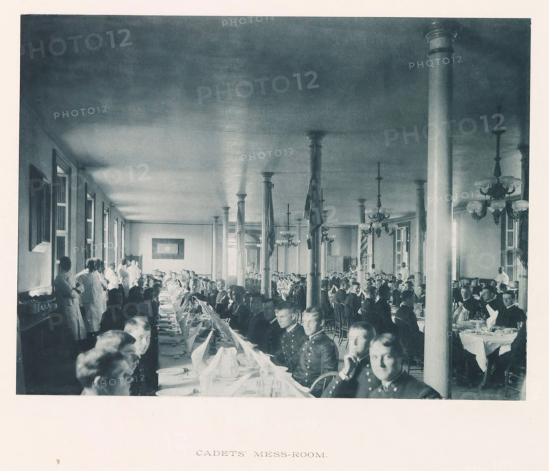 Cadet's mess-room, photograph, page, photographer: Edward H. Hart, United States Naval Academy, cliché maker: The Photo-gravure Co., New York (city), c. 1885 - in or before 1890, Edward H. Hart, The Photo-gravure Co., photographer, cliché maker, North America, Maryland, Annapolis, paper, collotype, photoprint, height 160 mm x width 215 mm, dining-room, officer, military academy, A large dining hall filled with long tables and many people seated along them, several uniformed individuals visible in the foreground, rows of standing columns running down the center of the room, chandeliers hanging from the ceiling, windows along the left wall, groups of staff in light-colored clothing standing along the left side, folded napkins and place settings on the tables, framed pictures on the walls, and flags hanging from some columns.