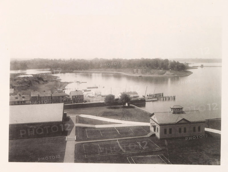 Looking north from tower, photograph, page, photographer: Edward H. Hart, United States Naval Academy, cliché maker: The Photo-gravure Co., New York (city), c. 1885 - in or before 1890, North America, Maryland, Annapolis, paper, collotype, photoprint, height 167 mm x width 238 mm, gulf, bay, A sepia-toned aerial view shows a waterfront scene with calm water, a curved shoreline lined with trees, and a small peninsula with a grassy slope. Several piers and docks extend into the water with small sailboats and rowboats moored nearby. A row of low buildings with pitched roofs sits along the near shore, and a larger building with a cupola and a facade facing the water occupies the foreground beside paved walkways and marked rectangular courts. Paths and grassy areas surround the buildings, and a distant treeline and a long bridge are visible along the horizon.
