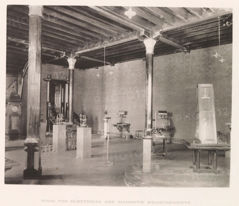 Room for electrical and magnetic measurements, photograph, page, photographer: Edward H. Hart, United States Naval Academy, cliché maker: The Photo-gravure Co., New York (city), c. 1885 - in or before 1890, North America, Maryland, Annapolis, paper, collotype, photoprint, height 185 mm x width 233 mm, laboratory, research room, military academy, A large interior space with exposed wooden ceiling beams supported by square and round columns with decorative capitals, brick walls, hanging light fixtures, several worktables and pedestals holding mechanical instruments and machines, a tall wooden cabinet leaning against a wall, stairs and a doorway partially visible in the background, and various cables and small equipment on the floor.