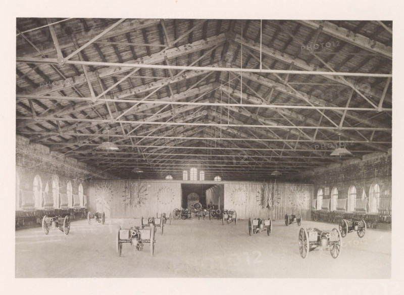 Armory, photograph, page, photographer: Edward H. Hart, United States Naval Academy, cliché maker: The Photo-gravure Co., New York (city), c. 1885 - in or before 1890, North America, Maryland, Annapolis, paper, collotype, photoprint, height 151 mm x width 219 mm, firearms: cannon, ammunition store, arsenal, A large interior space with a high wooden roof supported by exposed beams and trusses, hanging light fixtures suspended from the ceiling, brick side walls with a row of arched windows on each side, wooden flooring, a wide central doorway at the far wall flanked by decorative radial arrangements mounted on the wall, and several wheeled cannons or artillery pieces arranged across the floor.