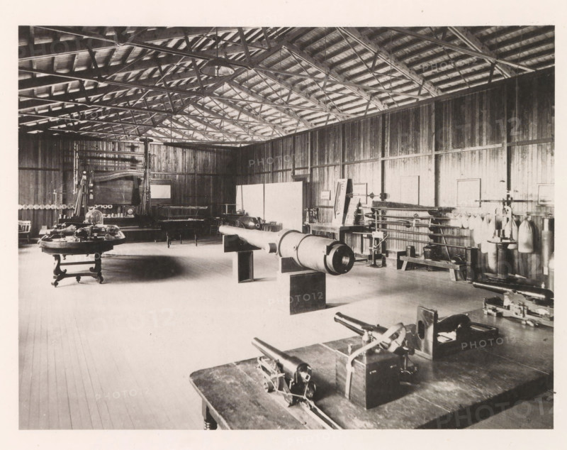Torpedo model room, photograph, page, photographer: Edward H. Hart, United States Naval Academy, cliché maker: The Photo-gravure Co., New York (city), c. 1885 - in or before 1890, North America, Maryland, Annapolis, paper, collotype, photoprint, height 176 mm x width 234 mm, firearms: cannon, ammunition store, arsenal, A wide interior view of a large wooden room with exposed roof trusses and rafters, wooden plank walls and a wooden floor, containing various pieces of equipment and furniture arranged across the space, including a long cylindrical object mounted on supports near the center, several tables with mechanical devices on them in the foreground and left side, a tall framed apparatus against the back wall, shelving and racks holding cylindrical and bag-like items along the right wall, chairs and benches scattered around, and pendant lights hanging from the ceiling.