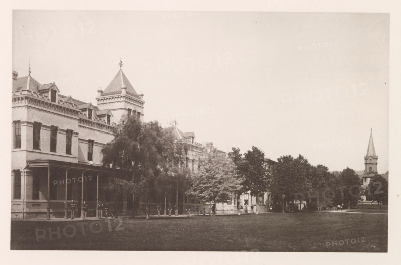 Porter Row, photograph, page, photographer: Edward H. Hart, United States Naval Academy, cliché maker: The Photo-gravure Co., New York (city), c. 1885 - in or before 1890, North America, Maryland, Annapolis, paper, collotype, photoprint, height 150 mm x width 237 mm, facade (of house or building), military academy, A wide sepia photograph showing the facade of a large brick building with decorative cornices, dormer windows, and a steep pyramidal tower topped with a finial, a covered porch with columns along its ground floor, and a row of trees partially obscuring the lower part of the building; several people stand or sit near the porch and along the sidewalk beneath the trees; a grassy lawn occupies the foreground, and to the right a church steeple rises above more trees with a small carriage visible near its base.
