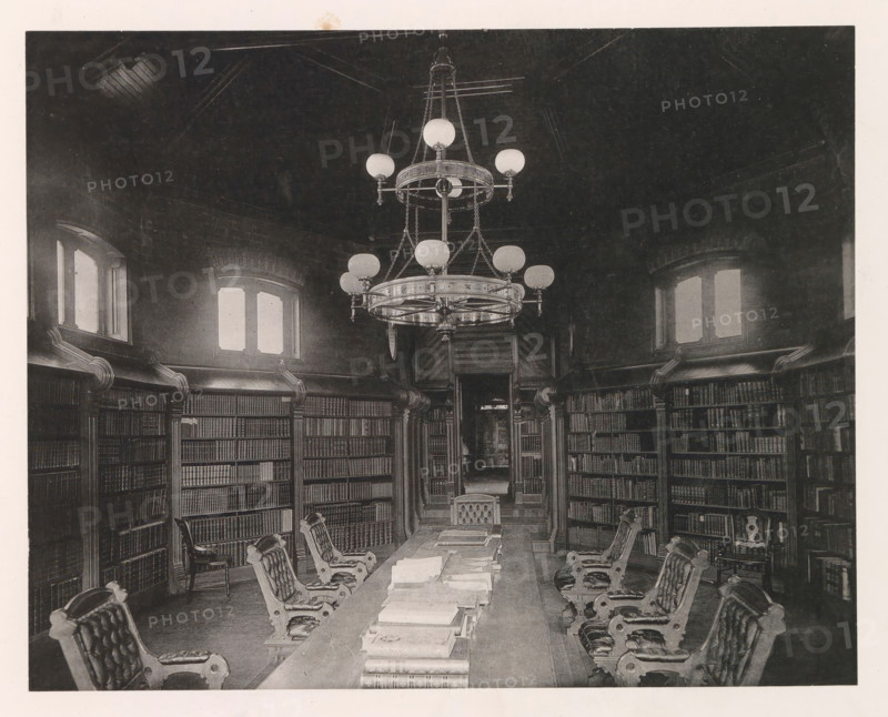 Chapel, photograph, page, photographer: Edward H. Hart, United States Naval Academy, cliché maker: The Photo-gravure Co., New York (city), c. 1885 - in or before 1890, North America, Maryland, Annapolis, paper, collotype, photoprint, height 177 mm x width 225 mm, public library, military academy, A high-ceilinged room with tall paneled bookshelves lining curved walls, several arched windows with light filtering through, a long central table covered with stacked books and papers, ornate upholstered chairs with carved wooden frames positioned along both sides of the table, a large multi-tiered chandelier hanging from the ceiling with spherical light fixtures and decorative chains, carved wooden columns and molding around the shelves, and a doorway visible at the far end leading to a darker interior space.