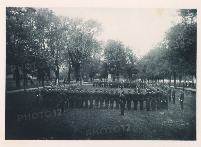 General muster, photograph, page, photographer: Edward H. Hart, United States Naval Academy, cliché maker: The Photo-gravure Co., New York (city), c. 1885 - in or before 1890, North America, Maryland, Annapolis, paper, collotype, photoprint, height 165 mm x width 240 mm, commander-in-chief, general, marshal, military academy, A wide outdoor scene showing a formal formation of uniformed individuals standing in concentric arcs on a grassy area surrounded by tall trees, with a central open space and an obelisk-like monument visible among the trees in the midground, pathways and scattered figures along the tree line, and a pale sky above.
