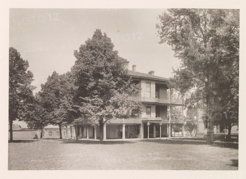 Sick-quarters, photograph, page, photographer: Edward H. Hart, United States Naval Academy, cliché maker: The Photo-gravure Co., New York (city), c. 1885 - in or before 1890, North America, Maryland, Annapolis, paper, collotype, photoprint, height 165 mm x width 236 mm, hospital, facade (of house or building), A large two-story building with a wraparound porch and ornate railing sits partially obscured by tall leafy trees in a grassy open area, with columns supporting the porch and a smaller covered entry at ground level; the building has tall windows and chimneys visible above the roof, and additional trees and parts of other brick structures are visible in the background.