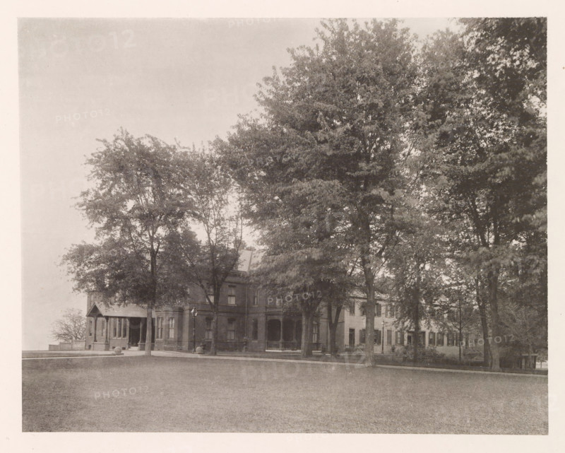 Superintendent's residence, photograph, page, photographer: Edward H. Hart, United States Naval Academy, cliché maker: The Photo-gravure Co., New York (city), c. 1885 - in or before 1890, Edward H. Hart, The Photo-gravure Co., photographer, cliché maker, North America, Maryland, Annapolis, paper, collotype, photoprint, height 185 mm x width 233 mm, housing, facade (of house or building), military academy, A wide lawn in the foreground leads to a row of large trees with dense canopies that dominate the middle of the image, partially obscuring a multi-story brick building with a porch, columns, and multiple windows; a smaller gabled entrance and a street lamp are visible near the building, and the scene is framed by a light border around the photograph.