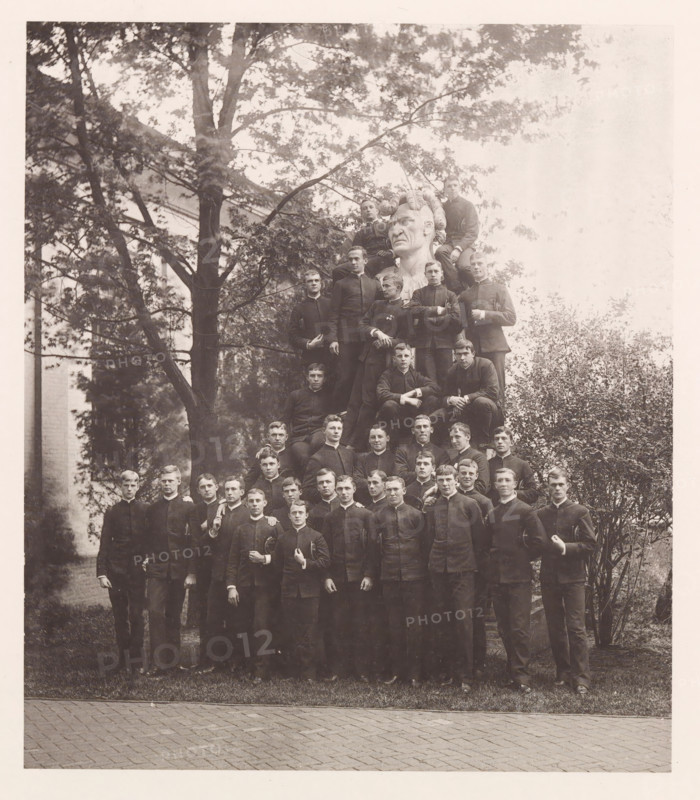 Class of '88, photograph, page, photographer: Edward H. Hart, United States Naval Academy, cliché maker: The Photo-gravure Co., New York (city), 1888, Edward H. Hart, The Photo-gravure Co., photographer, cliché maker, North America, Maryland, Annapolis, paper, collotype, photoprint, height 215 mm x width 185 mm, sculpture, officer, anonymous historical persons depicted in a group, in a group-portrait, A sepia-toned photograph shows a large group of young men arranged in rows outdoors, many wearing dark uniforms with high collars and holding hats or caps; some stand on the ground while others sit or perch higher, forming a pyramid shape around a large carved bust with a stern expression and stylized hair near the top center; behind them are tall trees with leafy branches and partial views of a building, and the foreground shows grass edging a paved surface.