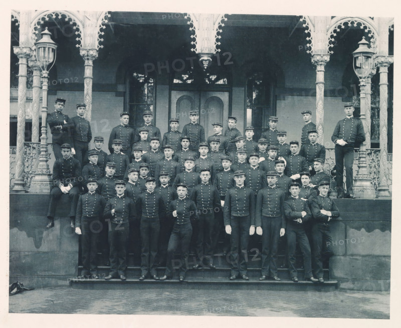 Class of '89, photograph, page, photographer: Edward H. Hart, United States Naval Academy, cliché maker: The Photo-gravure Co., New York (city), 1889, Edward H. Hart, The Photo-gravure Co., photographer, cliché maker, North America, Maryland, Annapolis, paper, collotype, photoprint, height 180 mm x width 228 mm, officer, anonymous historical persons depicted in a group, in a group-portrait, A large group of uniformed men posed on the steps of a building with ornate columns and arched decorative trim, many wearing dark buttoned jackets and peaked caps, some wearing white gloves, arranged in tiered rows across the stairs and landing, flanked by two decorative lampposts and carved pillars, with tall arched doorways and windows visible behind them and a stone base and patterned pavement in the foreground.