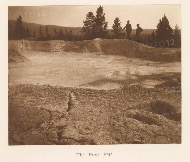 The paint pots, page, photomechanical print, photographer: Frank Jay Haynes, Yellowstone National Park, cliché maker: The Photo-gravure Co., New York (city), c. 1882 - in or before 1887, North America, paper, photoaquatint, height 170 mm x width 211 mm, pond, pool, hot spring, geyser, Fountain Paint Pot, A sepia-toned landscape photograph showing a rough, textured foreground of cracked and uneven ground leading to a flatter, lighter expanse and a low ridge; silhouettes of tall trees line the ridge against a pale sky, and three figures in dark clothing and hats stand near the ridge edge with two more distant tree silhouettes beyond.