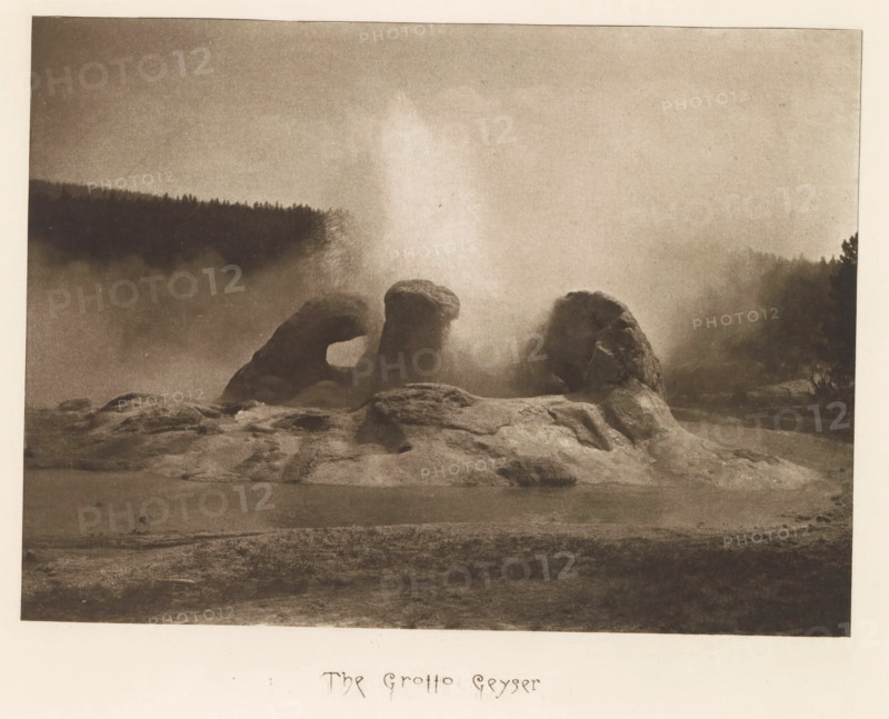 The Grotto Geyser, page, photomechanical print, photographer: Frank Jay Haynes, Yellowstone National Park, cliché maker: The Photo-gravure Co., New York (city), c. 1882 - in or before 1887, North America, paper, photoaquatint, height 153 mm x width 210 mm, hot spring, geyser, Grotto Geyser, A sepia-toned landscape photograph showing a rocky formation rising from a flat foreground, with three rounded rock masses positioned close together and a tall plume of mist or steam erupting between them; a dark treeline stretches across the background beneath a hazy sky, and diffuse mist hangs over the midground creating soft, blurred edges around the rocks and distant trees.