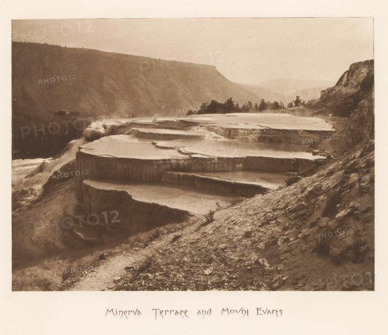 Minerva Terrace and Mount Evarts, page, photomechanical print, photographer: Frank Jay Haynes, Yellowstone National Park, cliché maker: The Photo-gravure Co., New York (city), c. 1882 - in or before 1887, North America, paper, photoaquatint, height 162 mm x width 216 mm, mountains, hot spring, geyser, Mount Everts, A sepia-toned landscape showing a series of broad terraced rock formations filled with shallow water cascading between levels, a steep rocky slope in the foreground with scattered small stones and sparse low vegetation, a distant ridge of hills fading into the background, a cluster of trees along the far terrace edge, and rocky cliffs rising on the right side of the image.