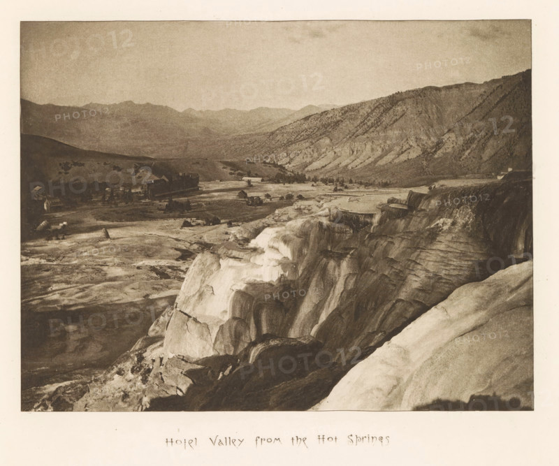 Hotel valley from the hot springs, page, photomechanical print, photographer: Frank Jay Haynes, Yellowstone National Park, cliché maker: The Photo-gravure Co., New York (city), c. 1882 - in or before 1887, North America, paper, photoaquatint, height 162 mm x width 216 mm, dale, valley, A wide sepia-toned landscape photograph showing a rocky foreground with terraces and smooth rock formations leading down toward a flat valley where small buildings and clustered trees are visible, beyond which low rolling hills and a range of rugged mountains extend across the background under a pale sky.