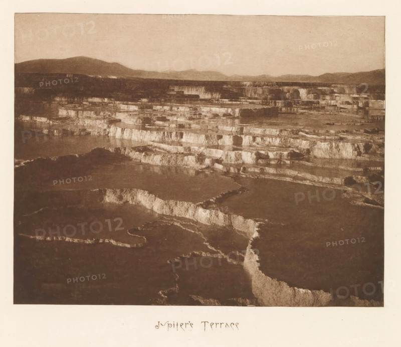 Jupiter's Terrace, page, photomechanical print, photographer: Frank Jay Haynes, Yellowstone National Park, cliché maker: The Photo-gravure Co., New York (city), c. 1882 - in or before 1887, Frank Jay Haynes, The Photo-gravure Co., photographer, cliché maker, North America, paper, photoaquatint, height 165 mm x width 213 mm, hot spring, geyser, A wide sepia-toned landscape image showing layered, stepped terraces with lighter horizontal edges and darker flat surfaces, stretching toward a distant horizon of low rounded hills beneath a pale sky, with irregular channels and textured surfaces visible across the terraces and a pale border surrounding the image.