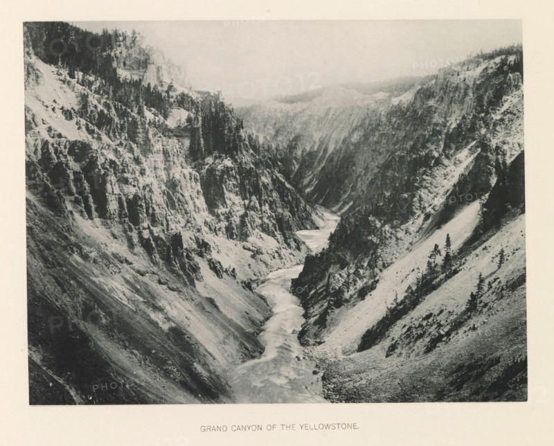 Grand canyon of the Yellowstone, page, photomechanical print, photographer: Frank Jay Haynes, Yellowstone National Park, cliché maker: The Photo-gravure Co., New York (city), c. 1886 - in or before 1891, North America, paper, collotype, photoprint, height 165 mm x width 215 mm, ravine, chasm, abyss, canyon, Grand Canyon (Yellowstone National Park), A wide grayscale photograph of a steep rocky canyon with rugged cliffs on both sides and a river winding through the canyon floor. The cliff faces show layered rock formations and scattered trees clinging to slopes and ledges, with denser stands of taller trees near the upper edges. The river appears lighter in tone against the darker canyon walls and flows from the distant background toward the foreground, with visible rapids and curving channels. The slopes beside the river vary from steep rocky ridges to smoother talus-covered areas dotted with small trees and shrubs. The upper background fades into a hazy sky above the distant ridgelines...