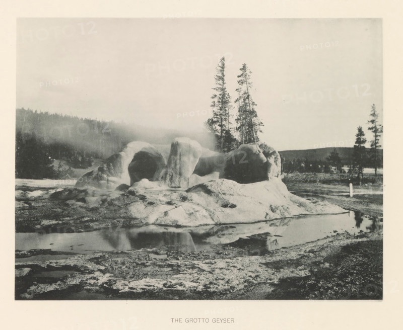 The Grotto Geyser, page, photomechanical print, photographer: Frank Jay Haynes, Yellowstone National Park, cliché maker: The Photo-gravure Co., New York (city), c. 1886 - in or before 1891, North America, paper, collotype, photoprint, height 165 mm x width 215 mm, hot spring, geyser, Grotto Geyser, A grayscale landscape shows a rounded rocky formation with smooth, bulbous surfaces and arch-like openings sitting beside a reflective pool of water, with the rock reflecting in the pool; behind the formation are tall, slender evergreen trees and a distant tree-covered hillside partially obscured by mist, while the foreground consists of rough, textured ground and patches of shallow water.