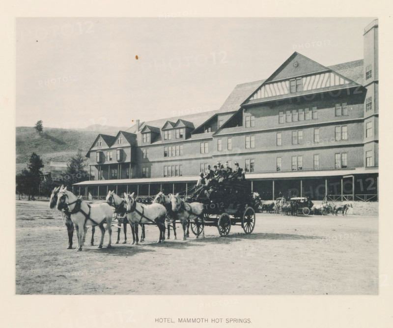 Hotel mammoth hot springs, page, photomechanical print, photographer: Frank Jay Haynes, Yellowstone National Park, cliché maker: The Photo-gravure Co., New York (city), c. 1886 - in or before 1891, North America, paper, collotype, photoprint, height 164 mm x width 216 mm, hotel, hostelry, inn, four-wheeled, animal-drawn vehicle, eg: cab, carriage, coach, façade (of house or building), A large multi-story wooden hotel with a long covered porch and multiple dormer windows fills the right and center of the scene, with a hillside and scattered trees visible behind it on the left; in front of the building a team of horses harnessed to a wagon stands in the foreground, several people seated on the wagon and others nearby, additional carriages and horses lined up along the porch, and the ground in front appearing as an open dirt area.