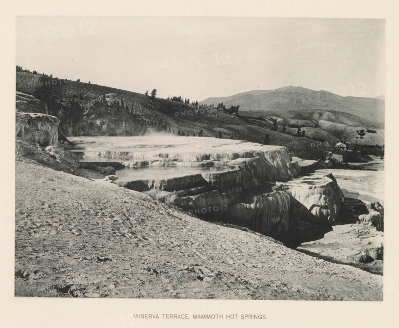 Minerva terrace, Mammoth hot springs, page, photomechanical print, photographer: Frank Jay Haynes, Yellowstone National Park, cliché maker: The Photo-gravure Co., New York (city), c. 1886 - in or before 1891, North America, paper, collotype, photoprint, height 164 mm x width 216 mm, hot spring, geyser, A wide landscape photograph showing layered mineral terraces with smooth, rounded edges and shallow pools, a steep rocky foreground slope leading down to the terraces, steam rising gently from some pools, a tree-covered hillside in the midground with scattered trees along the ridge, distant rolling mountains under a pale sky, and a small cluster of buildings near the right edge of the terraces.