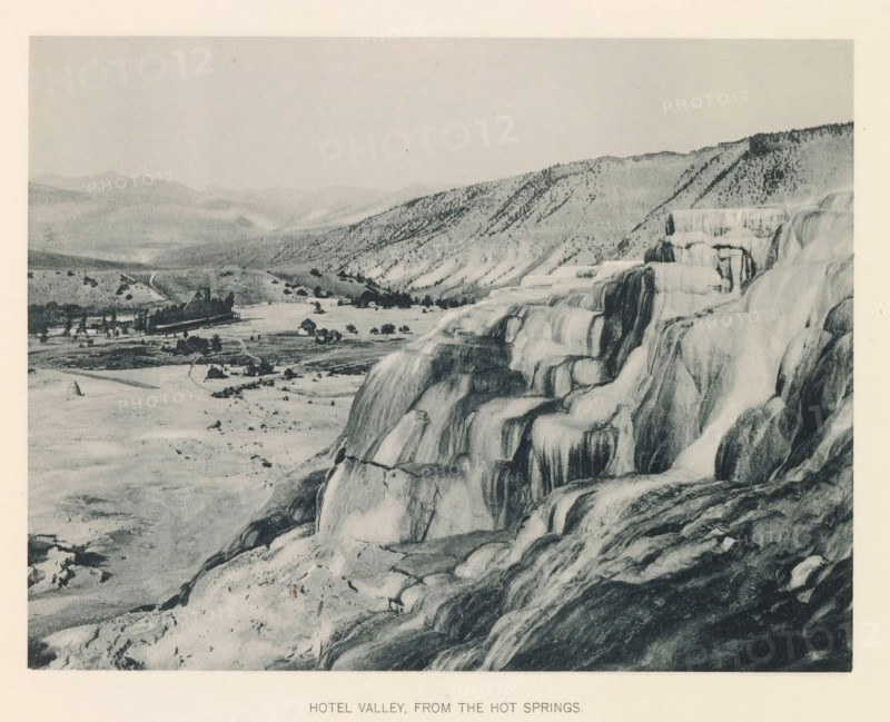 Hotel valley, from the hot springs, page, photomechanical print, photographer: Frank Jay Haynes, Yellowstone National Park, cliché maker: The Photo-gravure Co., New York (city), c. 1886 - in or before 1891, North America, paper, collotype, photoprint, height 165 mm x width 215 mm, dale, valley, hot spring, geyser, A wide landscape shows tiered mineral terraces with smooth rounded ledges and cascading surfaces in the foreground, their textures ranging from light to dark tones; beyond the terraces a flat valley contains scattered small buildings and a larger complex with peaked roofs, surrounded by open land and sparse trees; rolling hills and ridges rise in the background with scattered vegetation on their slopes, and a pale sky fills the upper portion of the scene.
