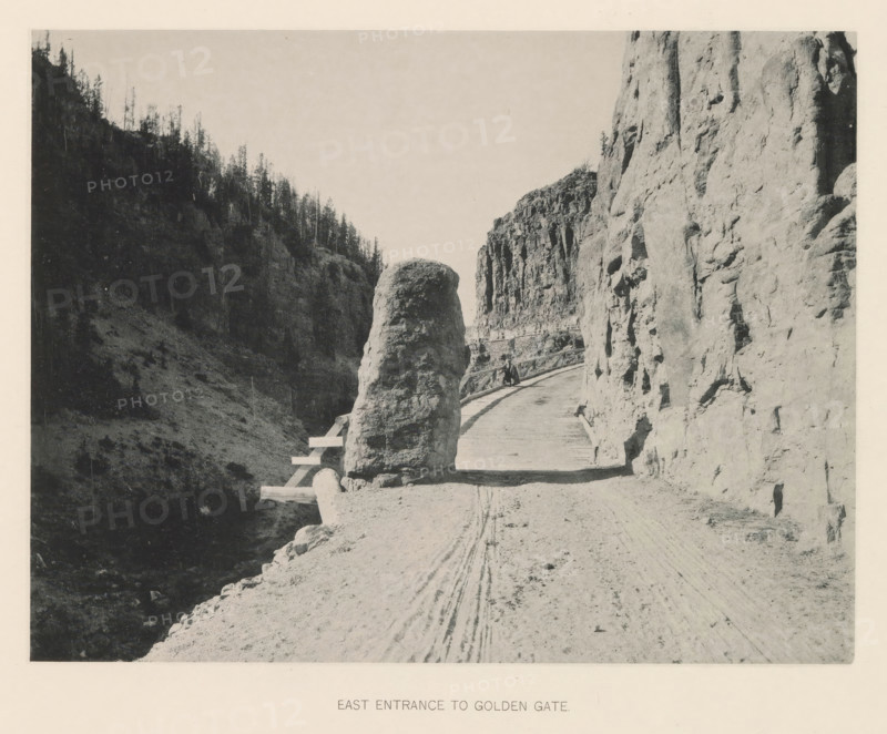 East entrance to Golden Gate, page, photomechanical print, photographer: Frank Jay Haynes, Yellowstone National Park, cliché maker: The Photo-gravure Co., New York (city), c. 1886 - in or before 1891, North America, paper, collotype, photoprint, height 165 mm x width 215 mm, public road, A narrow dirt road curves between steep rock walls with a wooden railing along its outer edge, a large rounded boulder sits near the road’s inner side, tall rocky cliffs rise on either side with rough textured surfaces, a figure appears seated on the railing partway along the road, tire tracks are visible in the dirt, distant trees grow on the ridge of the left cliff, and the sky above is clear.
