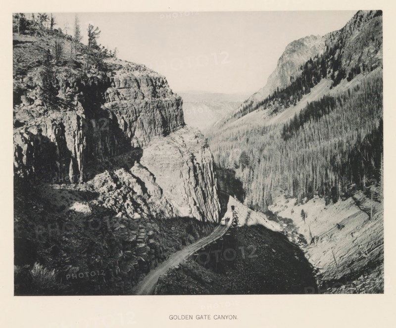 Golden Gate Canyon, page, photomechanical print, photographer: Frank Jay Haynes, Yellowstone National Park, cliché maker: The Photo-gravure Co., New York (city), c. 1886 - in or before 1891, North America, paper, collotype, photoprint, height 165 mm x width 215 mm, ravine, chasm, abyss, canyon, A black and white image showing a rugged canyon with steep rock cliffs on one side and a densely forested slope on the other, a narrow winding road or wooden trestle running along the canyon edge, sparse trees and shrubs on the rocky ledges, and distant layered hills visible through the canyon opening.