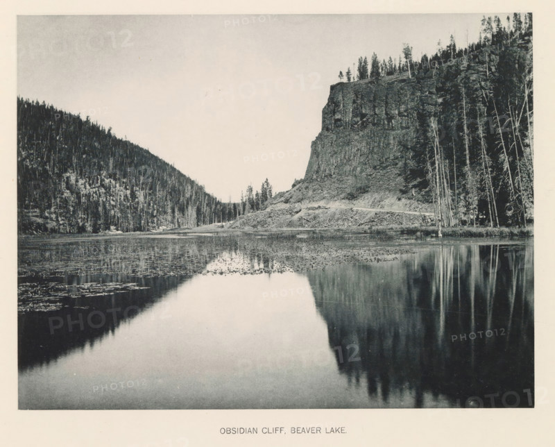 Obsidian cliff, Beaver Lake, page, photomechanical print, photographer: Frank Jay Haynes, Yellowstone National Park, cliché maker: The Photo-gravure Co., New York (city), c. 1886 - in or before 1891, North America, paper, collotype, photoprint, height 165 mm x width 215 mm, ravine, chasm, abyss, canyon, A wide black-and-white photograph shows a calm lake in the foreground reflecting the cliff and trees along its shore, with the water surface dotted with patches of vegetation and subtle ripples; to the right a tall rocky cliff rises from a sloping shoreline, topped by a line of trees and bordered by a stand of tall, thin trunks along the water's edge, while to the left a densely forested hillside slopes down to meet the shore, and a pale sky fills the upper area above the landscape.