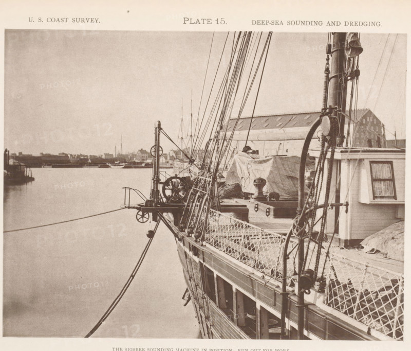 View of the Sigsbee sounding machine at the George S. Blake, page, photomechanical print, inscription: 'The Sigsbee sounding machine in position: run out for work.', number: 'Plate 15.', name: 'Heliotype Printing Co., 220 Devonshire St., Boston.', United States of America, printer (person): Heliotype Printing Company, Boston (Massachusetts), c. 1870 - in or before 1880, printer (person), North America, Massachusetts, paper, collotype, photoprint, height 165 mm x width 215 mm, steamship, motorship, machines, parts of machines, tools and appliances, fathoming, sounding, A sepia-toned photograph showing the side and deck of a sailing vessel with ropes, rigging, pulleys, and netted railings, a covered area on deck with tarpaulin and mechanical winches, a tall mast with additional rigging and a coiled rope, a small cabin window with curtains, and a calm body of water alongside a distant shoreline with low buildings and moored boats visible on the horizon.