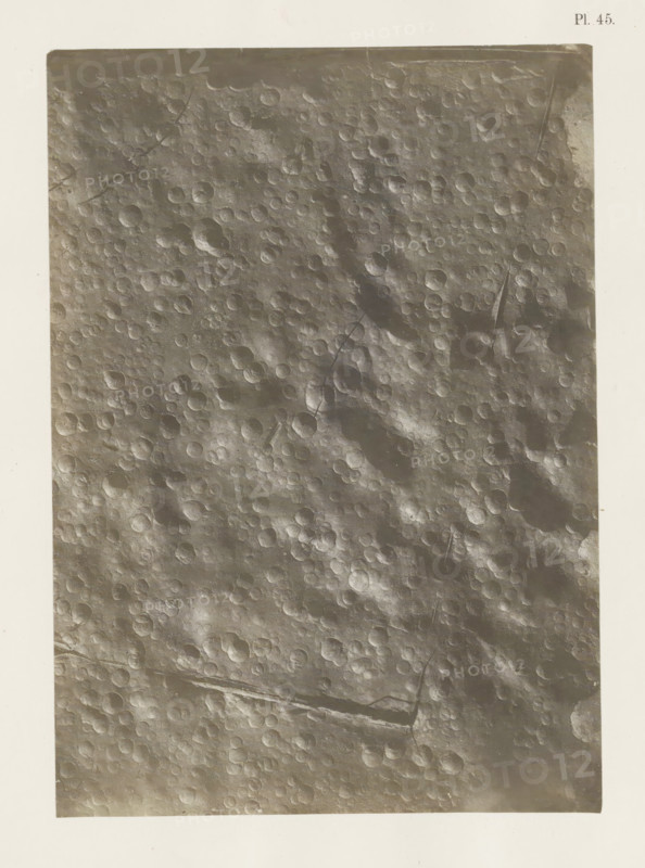 Print of raindrops, photograph, page, number: 'Pl. 45.', United States of America, c. 1850 - in or before 1861, photographer, North America, paper, salted paper print, height 201 mm x width 143 mm, Photo in section at the back of the book., rain, foot-print, track, A rectangular grayscale image bordered by a wide light margin shows a textured surface covered in many shallow circular depressions with raised rims, creating a pitted pattern across the frame; the surface varies in tone with lighter and darker patches, subtle ridges and elongated grooves cross some areas, and there are thin linear scratches and faint smudges visible against the otherwise rough, cratered texture.