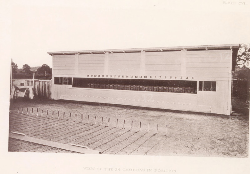 View of the 24 cameras in position, Setup with photo cameras to capture a horse in motion, page, photomechanical print, number: 'Plate_CVI.', photographer: Eadweard Muybridge, Stanford, cliché maker: anonymous, c. 1878 - in or before 1882, clichémaker, paper, photoaquatint, height 133 mm x width 217 mm, taking photographs - CC - out of doors, photographic apparatus, A long rectangular wooden structure with a slanted roof stands horizontally across the scene; its front face has a series of dark rectangular openings along a midline and a row of small square compartments beneath a sheltered overhang, with vertical supports dividing the panels. In front of the structure is an open yard with a boarded surface in the foreground featuring evenly spaced slender upright posts, and a patch of bare ground between the boards and the building. To the left of the structure there is a fenced area with a simple wooden frame and leaning planks, and low distant trees and shrubbery are visible along the ...