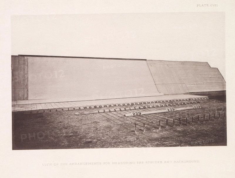 Arrangement with pedometers for photographing a horse in motion, View of the arrangement for measuring the strides and background, page, photomechanical print, number: 'Plate_CVII.', photographer: Eadweard Muybridge, Stanford, cliché maker: anonymous, c. 1878 - in or before 1882, clichémaker, paper, photoaquatint, height 138 mm x width 221 mm, taking photographs - CC - out of doors, A sepia-toned photograph showing a large flat vertical surface occupying the upper portion of the image, with a sloped wooden or paneled section on the right and a vertical framed edge on the left, a narrow horizontal ledge running along the base of the vertical surface, and a wooden platform in front of it marked with regularly spaced rectangular panels; in the foreground a textured ground area with a series of thin vertical stakes casting shadows and a few rectangular markers placed on the ground, all set within a pale border.
