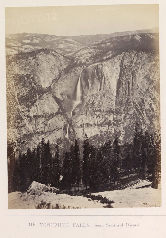 The Yosemite Falls, from Sentinel Dome, photograph, page, number: 'XI.', photographer: Carleton E. Watkins, Yosemite Valley, c. 1860 - in or before 1868, North America, California, paper, albumen print, height 206 mm x width 156 mm, waterfall, mountains, dale, valley, Yosemite National Park, Yosemitewaterval, A sepia-toned vertical photograph showing a steep rocky valley with a tall, narrow waterfall cascading down a cliff face toward a lower drop, surrounding rock formations with textured surfaces and scattered trees on the slopes, a dense stand of conifer trees in the foreground on a snowy slope, a distant ridgeline of rounded hills under a pale sky, and a light border around the image.