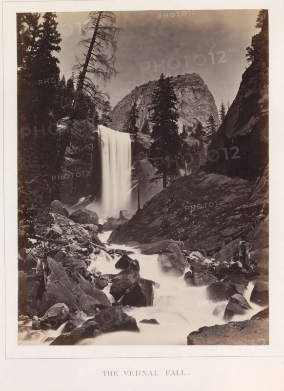 View of Vernal Falls in Yosemite Valley, The Vernal Fall, photograph, page, number: 'XVI.', photographer: Carleton E. Watkins, Yosemite Valley, c. 1860 - in or before 1868, North America, California, paper, albumen print, height 208 mm x width 155 mm, waterfall, mountains, rock-formations, dale, valley, Vernalwaterval, A tall waterfall cascades from a rocky ledge into a turbulent stream that flows over numerous boulders and rocks, with mist rising where the water impacts; on either side of the falls are steep rock faces and a variety of conifer trees, some tall and full, others bare and leaning, with a large rounded granite cliff visible in the background beneath a cloudy sky.