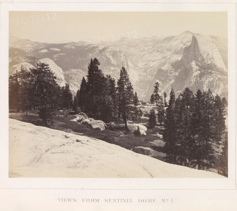 View of the Yosemite Valley from Sentinel Dome, Views from Sentinel Dome No. 1, photograph, page, number: 'XIX.', photographer: Carleton E. Watkins, Yosemite Valley, c. 1860 - in or before 1868, North America, California, paper, albumen print, height 156 mm x width 206 mm, mountains, rock-formations, dale, valley, A wide sepia-toned landscape photograph showing a mountainous scene with a foreground of a rounded rocky slope and scattered boulders, a midground populated by clusters of tall evergreen trees and open grassy patches, and a distant backdrop of layered mountain ridges and steep granite cliffs with a prominent sheer rock face rising on the right side, all under a pale sky.