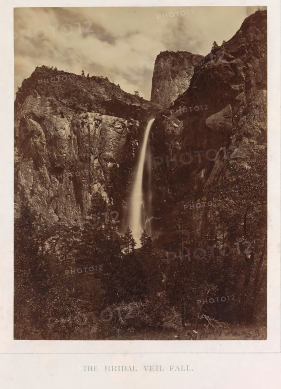 View of Bridalveil Falls in Yosemite Valley, The Bridal Veil Fall, photograph, page, number: 'II.', photographer: Carleton E. Watkins, Yosemite Valley, c. 1860 - in or before 1868, North America, California, paper, albumen print, height 204 mm x width 155 mm, waterfall, mountains, dale, valley, Bridalveilwaterval, A tall, narrow waterfall cascades down a steep rock face between rugged cliffs, the water appearing smooth and slightly blurred as it falls into a darker, wooded area below; surrounding the waterfall are high, jagged rock formations with visible striations and scattered trees along the ridgelines, and above them a cloudy sky with soft, diffuse light.