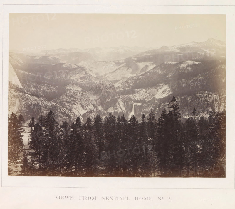 View of the Yosemite Valley from Sentinel Dome, Views from Sentinel Dome No. 2, photograph, page, number: 'XX.', photographer: Carleton E. Watkins, Yosemite Valley, c. 1860 - in or before 1868, North America, California, paper, albumen print, height 156 mm x width 206 mm, mountains, rock-formations, dale, valley, A wide sepia-toned landscape photograph showing a mountainous valley scene with snow-dusted peaks and rounded granite domes receding into a hazy sky, steep rocky cliffs and ridges forming layered slopes across the middle distance, a waterfall cascading down a sheer rock face near the center, and a dense foreground of tall evergreen trees partially covering lower rock formations.
