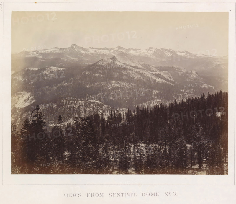 View of the Yosemite Valley from Sentinel Dome, Views from Sentinel Dome No. 3, photograph, page, number: 'XXI.', photographer: Carleton E. Watkins, Yosemite Valley, c. 1860 - in or before 1868, North America, California, paper, albumen print, height 155 mm x width 207 mm, mountains, rock-formations, dale, valley, A wide, sepia-toned landscape photograph showing layered mountain ridges receding into the distance under a pale sky, with snow patches on higher slopes and darker tree-covered lower slopes; the foreground contains a dense forest of tall conifer trees with snow on branches and a textured, uneven ground; midground domes and rounded granite outcrops rise above the tree line, and distant jagged peaks with snow traces form a horizon line.