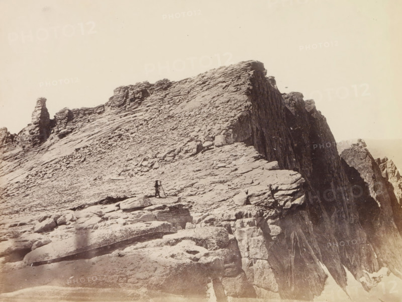 View of summit of Mount Hoffmann in the Yosemite Valley, Summit of Mt. Hoffmann, photograph, page, number: 'XXV.', photographer: W. Harris, Yosemite Valley, c. 1860 - in or before 1868, North America, California, paper, albumen print, height 157 mm x width 209 mm, mountain-top, peak, Mount Hoffmann, A sepia-toned photograph of a rocky mountain ridge with layered, jagged rock formations extending toward a steep cliff face, scattered boulders and slabs on the sloping foreground, a lone figure standing near a tripod on the rocky surface, and a tall, narrow rock outcrop visible farther along the ridge against a pale sky.