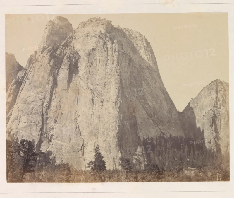 Cathedral Rock, near view from the front, View of the Middle Cathedral Rock in the Yosemite Valley, photograph, page, number: 'V.', photographer: Carleton E. Watkins, Yosemite Valley, c. 1860 - in or before 1868, North America, California, paper, albumen print, height 156 mm x width 206 mm, mountains, rock-formations, dale, valley, A sepia-toned photograph showing a steep, sheer rock face rising from a forested base, with rugged vertical cracks and ledges, lighter rock surfaces on sunlit faces and darker shaded areas creating depth, a few scattered trees and shrubs clinging near the top and along narrow terraces, a smaller rocky peak visible to the right, and a band of trees and vegetation at the bottom edge.