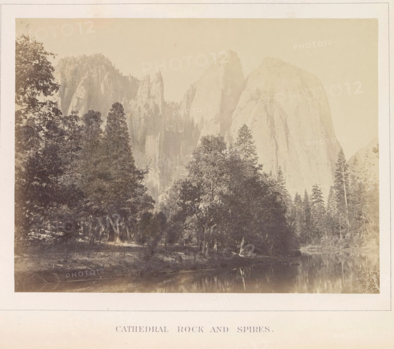 View of Middle Cathedral Rock and other spiers in the Yosemite Valley, Cathedral Rock ans Spires, photograph, page, number: 'VI.', photographer: Carleton E. Watkins, Yosemite Valley, c. 1860 - in or before 1868, North America, California, paper, albumen print, height 155 mm x width 206 mm, mountains, rock-formations, dale, valley, A sepia-toned landscape photograph showing tall rocky cliffs and spires rising behind a dense stand of trees, with a variety of tree silhouettes in the foreground and a calm reflective body of water along the lower edge, all framed by a pale border.