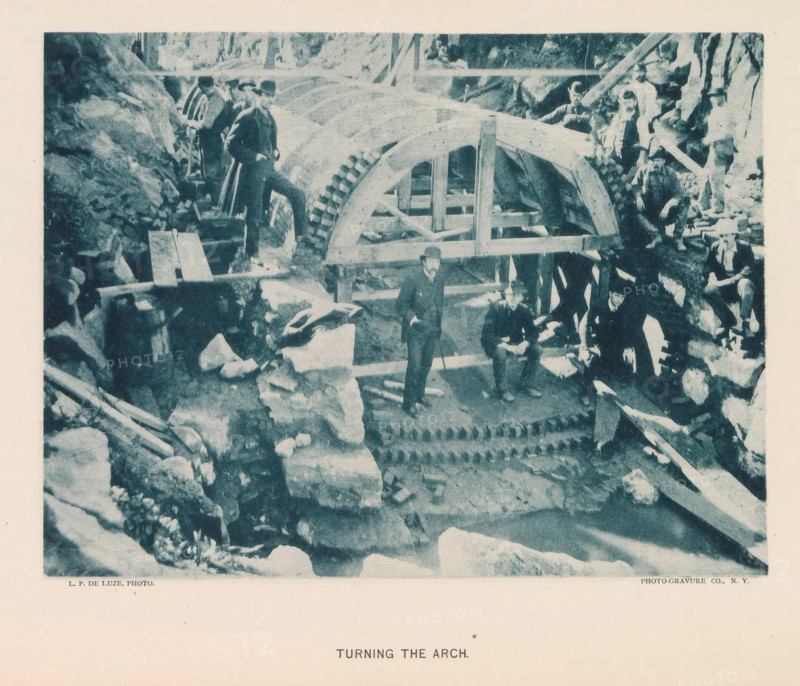Turning the arch, page, photomechanical print, photographer: Hall & Son, George P., New York, cliché maker: The Photo-gravure Co., c. 1883 - in or before 1887, North America, paper, collotype, photoprint, height 153 mm x width 202 mm, building-site, arch, archivolt, architecture, A monochrome photograph shows a group of men around a large wooden arch structure with visible gear-like teeth on one edge, set among rocky terrain and timber supports; some men stand on the arch or on surrounding rocks while others sit on beams, all wearing period clothing and hats, and various wooden planks, beams, and stone blocks are scattered around the worksite.