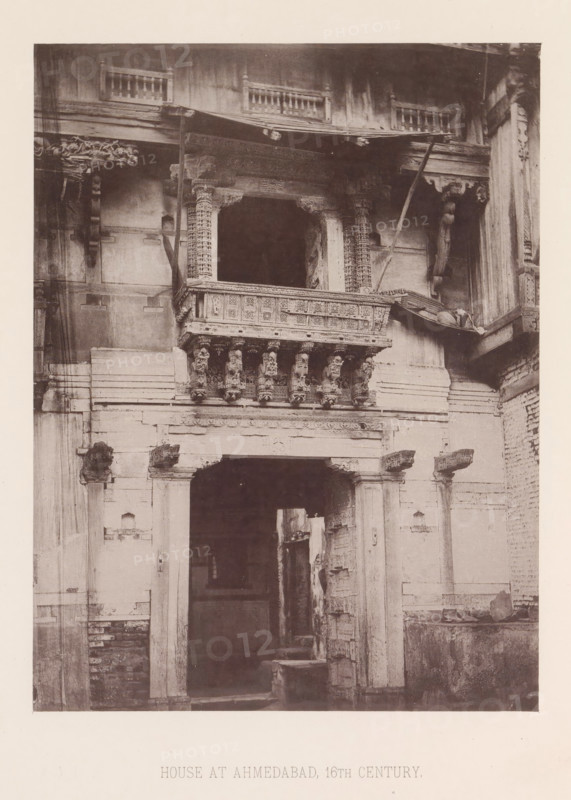 Façade of a house in Ahmedabad, Gujarat, India, page, photomechanical print, number: 'I.', title: 'HOUSE AT AHMEDABAD, 16TH CENTURY.', Ahmedabad (city), maker: Heliotype Printing Company, Boston (Massachusetts), c. 1875 - in or before 1885, photographer, North America, Massachusetts, paper, photoaquatint, height 230 mm x width 173 mm, urban housing, facade (of house or building), A sepia-toned photograph of a building facade showing an ornate balcony with carved pillars and decorative brackets above a large arched doorway. The balcony features intricate latticework panels and a row of sculpted corbels beneath its ledge. Flanking the doorway are weathered stone columns and remnants of carved details, with visible cracks and surface wear on the walls. Above the balcony is a wooden overhang with exposed beams and small balustrades along the upper level. The lower portion of the image shows the dark interior beyond the doorway and rough masonry at the base of the wall.