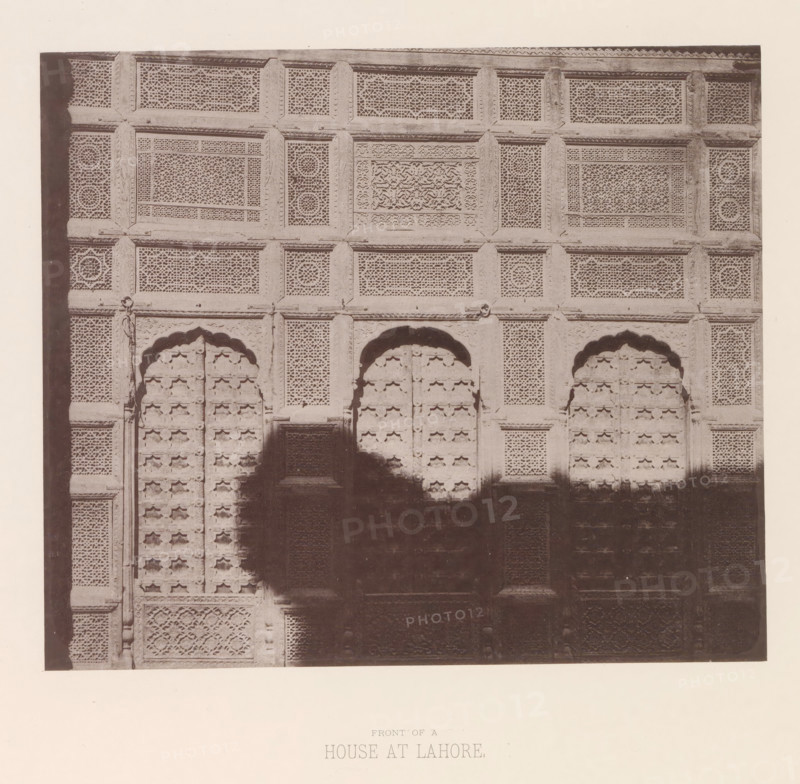 Façade of a house in Lahore, Punjab, Pakistan, page, photomechanical print, number: 'XI.', title: 'FRONT OF A, HOUSE AT LAHORE.', Lahore, maker: Heliotype Printing Company, Boston (Massachusetts), c. 1875 - in or before 1885, photographer, North America, Massachusetts, paper, photoaquatint, height 169 mm x width 198 mm, urban housing, facade (of house or building), A sepia photograph shows a large carved wooden or stone façade with multiple rectangular panels filled with intricate latticework and geometric patterns, framed by raised borders and carved vertical and horizontal beams; three arched recessed doorways with decorative molding are visible in the middle portion, each door set with repeating ornamental motifs, and shadowed areas obscure parts of the lower central section; the surface displays varied textures from finely pierced screens to carved relief panels, and the image is bordered by a plain margin.