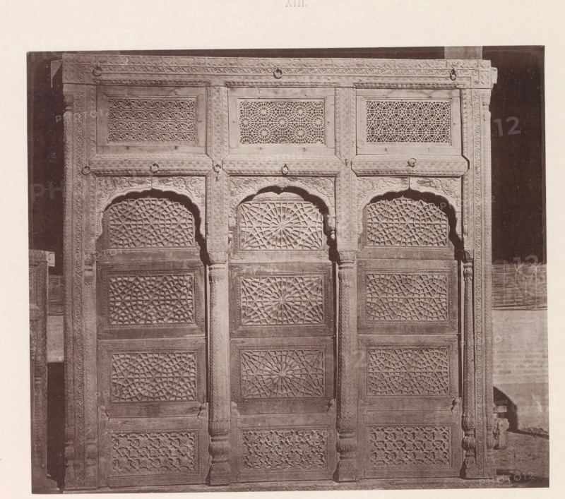 Window with geometrical ornaments in Lahore, Punjab, Pakistan, page, photomechanical print, title: 'WINDOW OF A, HOUSE AT LAHORE.', number: 'XIII.', Lahore, maker: Heliotype Printing Company, Boston (Massachusetts), c. 1875 - in or before 1885, photographer, North America, Massachusetts, paper, photoaquatint, height 177 mm x width 207 mm, ornament, geometric motifs, window, A carved wooden panel or door divided into vertical sections with repeating geometric lattice panels and ornate floral and rope-like border carvings, featuring arched recessed tops above central panels and fluted columns between sections, all shown in a monochrome photograph with a dark background and a light margin around the image.