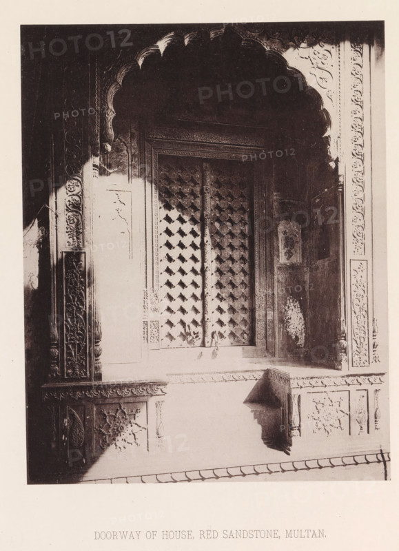 View of a doorway of a house in Multan, Punjab, Pakistan, page, photomechanical print, number: 'XIV.', title: 'DOORWAY OF HOUSE, RED SANDSTONE, MULTAN.', Multan, maker: Heliotype Printing Company, Boston (Massachusetts), c. 1875 - in or before 1885, photographer, North America, Massachusetts, paper, photoaquatint, height 221 mm x width 175 mm, closed door, A sepia-toned photograph showing a carved doorway with an ornate scalloped arch at the top and decorative panels along the sides. The central door appears to have a patterned lattice or grill with repeating geometric motifs, framed by intricately carved borders. The surrounding stonework features floral and vine reliefs, rosette medallions, and rectangular panels with carved details. Shadows fall across parts of the arch and lower wall, and there is a ledge or sill beneath the doorway with additional carved ornamentation. The image is bordered by a light margin.