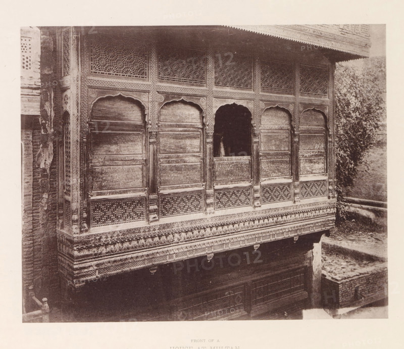 Façade of a house in Multan, Pubjab, Pakistan, page, photomechanical print, number: 'XV.', title: 'FRONT OF A, HOUSE AT MULTAN.', Multan, maker: Heliotype Printing Company, Boston (Massachusetts), c. 1875 - in or before 1885, photographer, North America, Massachusetts, paper, photoaquatint, height 187 mm x width 232 mm, urban housing, façade (of house or building), A sepia-toned photograph showing an ornate wooden façade with a row of arched openings framed by carved panels and decorative latticework above, intricate geometric and floral carvings along the columns and frieze, patterned balustrade and carved molding beneath the openings, weathered wood surfaces, a darker recessed central arch, brickwork visible at the left edge, and foliage or a tree at the right edge.