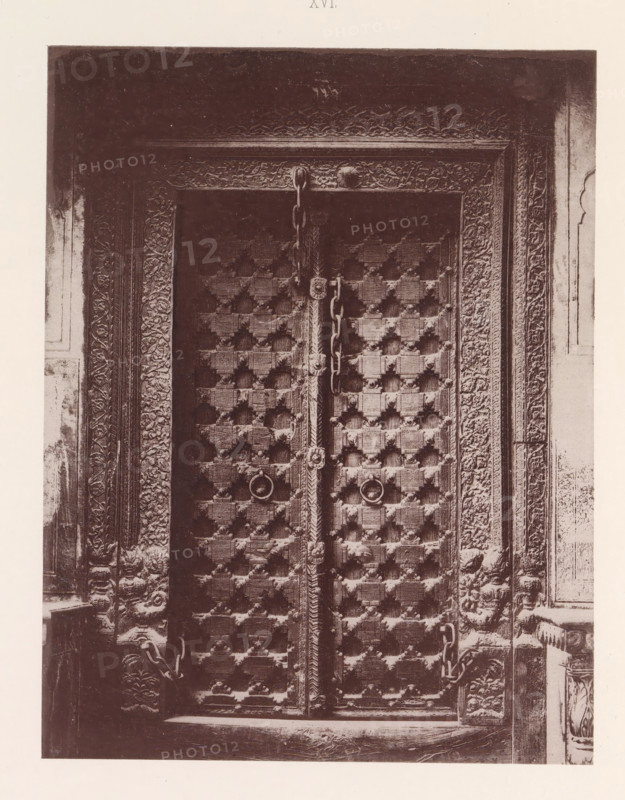 Door of a house in Multan, Punjab, Pakistan, page, photomechanical print, number: 'XVI.', title: 'DOOR OF HOUSE, MULTAN.', Multan, maker: Heliotype Printing Company, Boston (Massachusetts), c. 1875 - in or before 1885, photographer, North America, Massachusetts, paper, photoaquatint, height 221 mm x width 170 mm, closed door, A close view of an ornate wooden double door with a patterned grid of raised square studs and round metal ring handles, each door edged with carved vertical panels and a decorative central vertical strip; heavy metal chains hang across the doors and loop through anchors, and the surrounding frame is richly carved with floral and foliate motifs extending into the adjacent wall surfaces.
