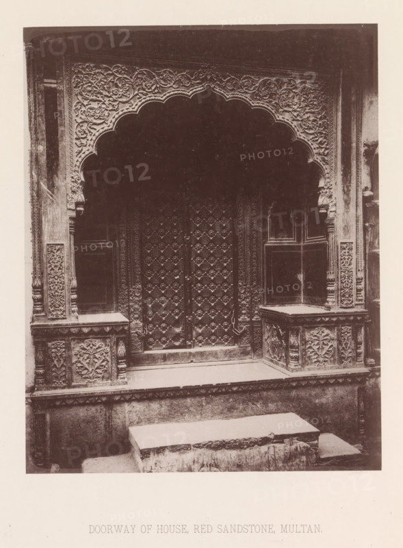 View of a doorway of a house in Multan, Punjab, Pakistan, page, photomechanical print, title: 'DOORWAY OF HOUSE, RED SANDSTONE, MULTAN.', number: 'XVIII.', Multan, maker: Heliotype Printing Company, Boston (Massachusetts), c. 1875 - in or before 1885, photographer, North America, Massachusetts, paper, photoaquatint, height 214 mm x width 170 mm, gate, entrance, closed door, A sepia-toned photograph of an ornate stone doorway featuring a scalloped arch with dense floral and vine carvings along its border, flanked by carved pilasters and panels with repeating floral motifs, a heavily decorated pair of wooden doors with geometric and floral relief patterns, raised stone platforms or benches on either side of the entrance with carved front panels, a low stone step leading up to the doorway, and weathered stone surfaces throughout.